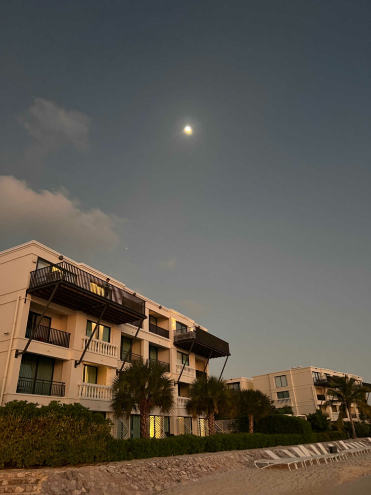 a view of the Curaçao Marriott Beach Resort taken from the beach. It is evening and there is a full moon over the resort