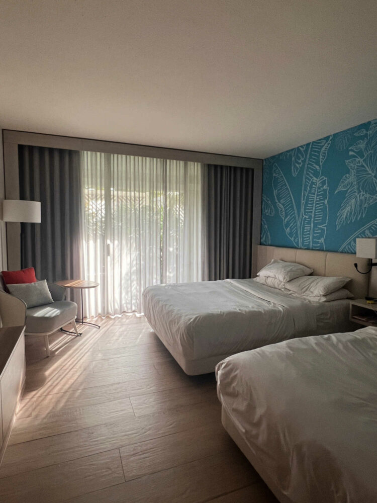 a view in a double queen garden room at the Curacao Marriott Beach Resort. Shows two queen beds, a sitting area, and patio doors opening to a private patio with tropical greenery beyond