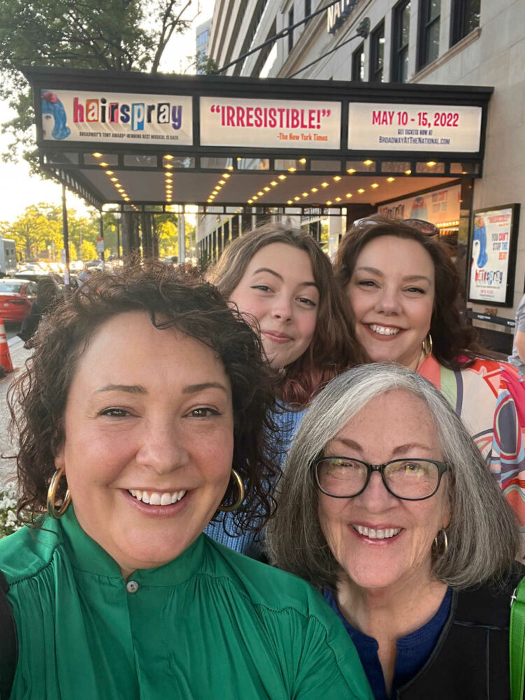 a mother, two middle aged daughters and a teen granddaughter are standing in front of the National Theater in DC. They are smiling and the theater marquee is featuring the musical Hairspray