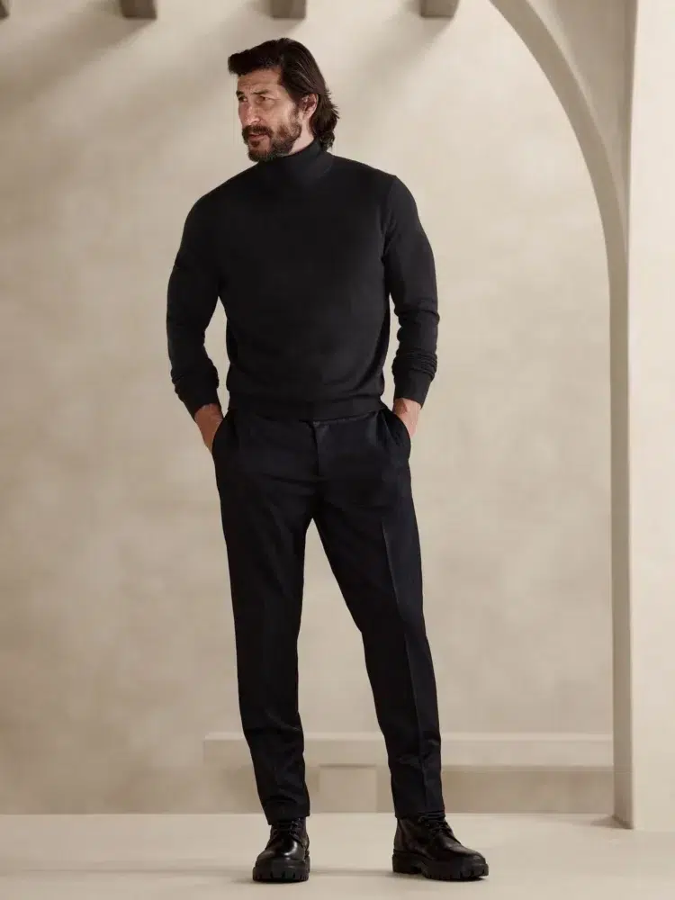 a man with brown hair wearing a black Banana Republic merino turtleneck with black trousers. His hands are in his pockets and he's look away from the camera