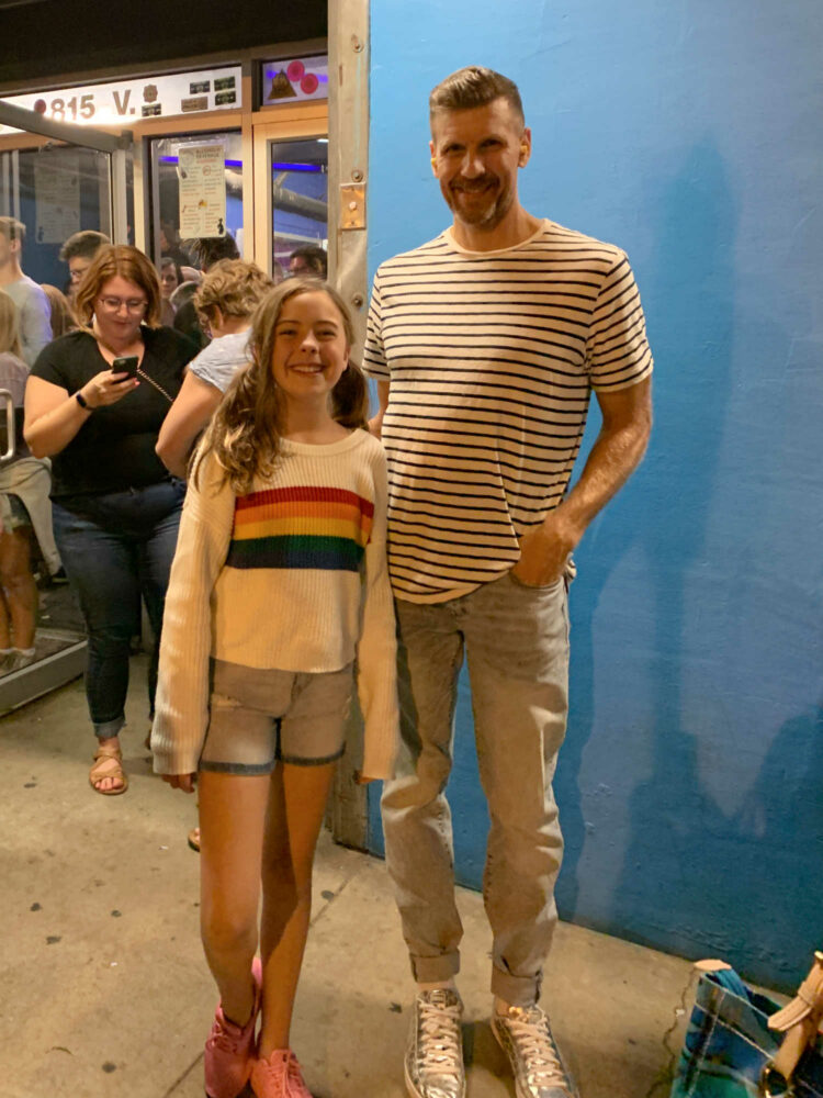 man in a white j. crew striped shirt and jeans standing next to a tween girl in a rainbow striped sweater and denim shorts. They are standing out side the 9:30 Club in Washington DC