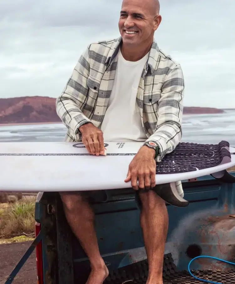 pro surfer Kelly Slater sitting on the back of a truck holding a surfboard. He is smiling and wearing an Outerknown blanket shirt in plaid open over a white t-shirt