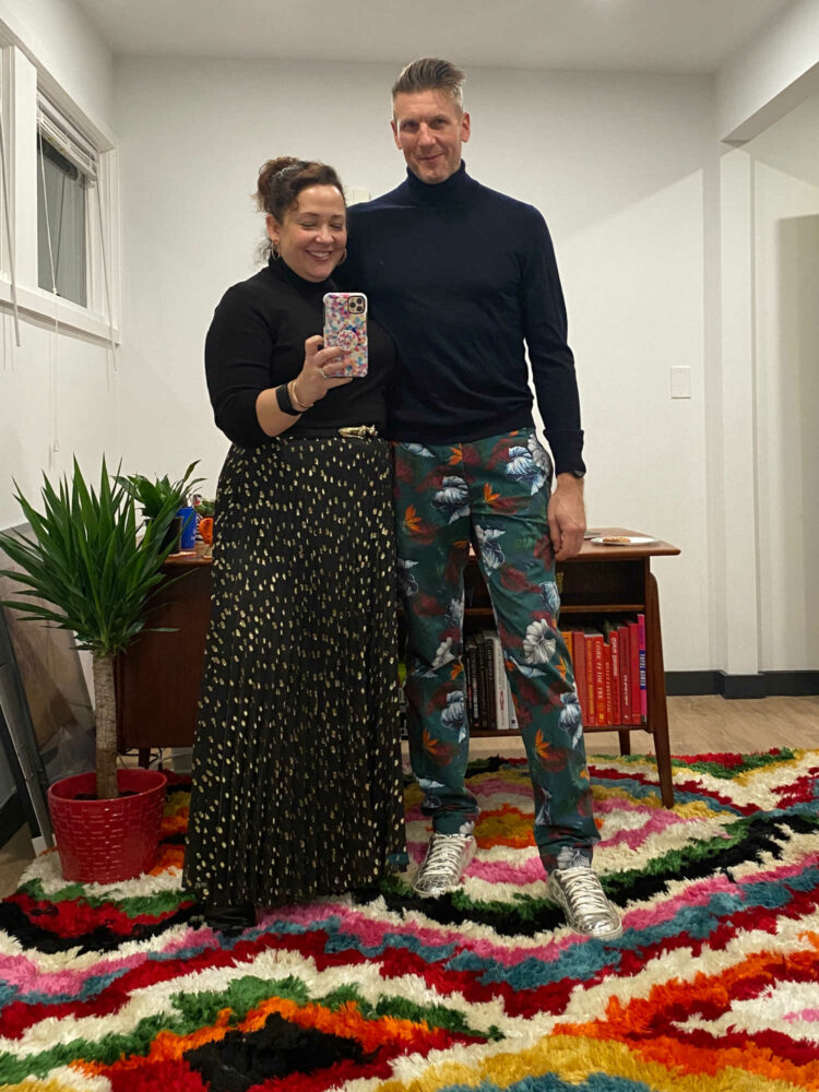 Alison and Karl Gary taking a mirror selfie. She is wearing a black turtleneck and black pleated maxi skirt with gold foil dots. He is in a navy Banana Republic merino turtleneck with navy floral chino pants.