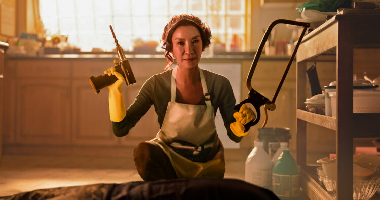 Scene from the Netflix series The Brothers Sun. Actress Michelle Yeoh is sitting on a kitchen floor holding a drill in one hand, a chainsaw in the other. She is wearing an apron and a scarf over her hair and a dead body is on the floor in front of her. She has a face of serenity.