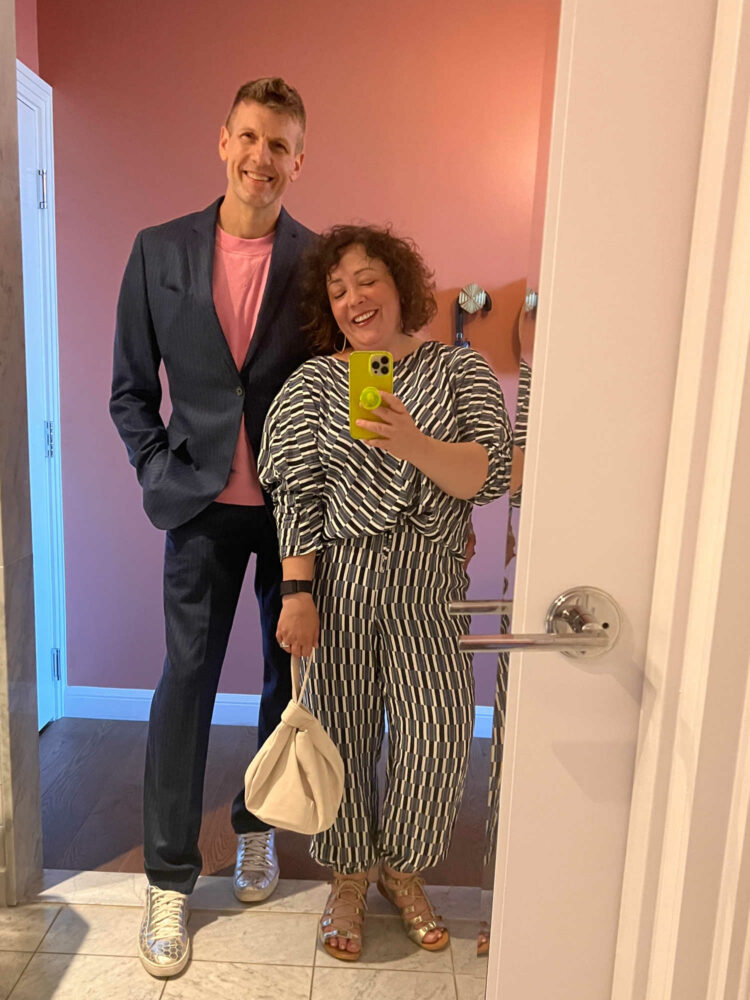 a man and woman taking a mirror selfie. The man is in a navy chalkstripe suit from Banana Republic with a pink t-shirt and silver Puma sneakers. The woman is in an M.M.LaFleur washable silk top and matching joggers and is holding an ivory handbag.