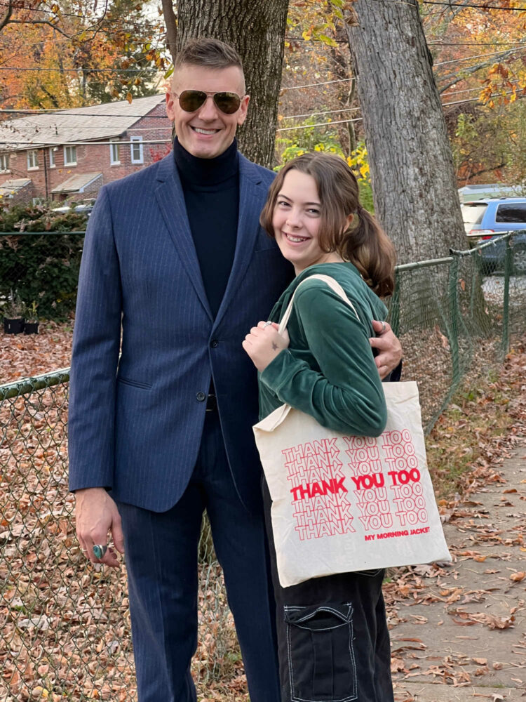 man in a navy Banana Republic pinstripe pantsuit with a navy turtleneck underneath. He is wearing aviator sunglasses and smiling at the camera. His arm is around a tween girl in a green velour hoodie and black jeans holding a canvas tote bag. She too is smiling at the camera.