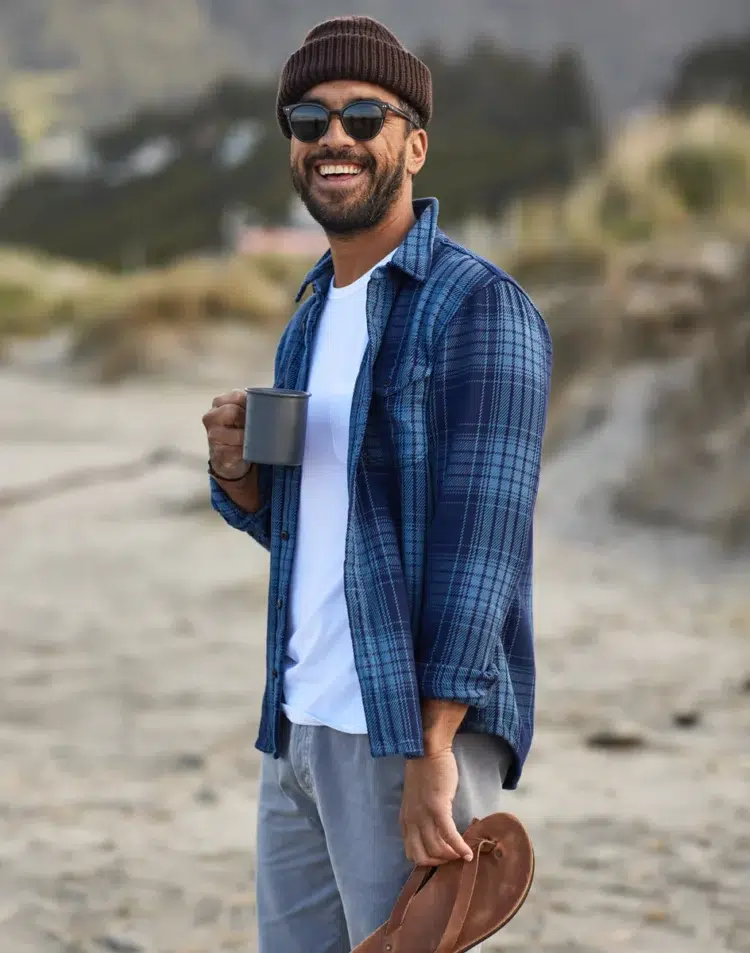 man on a beach wearing a ribbed beanie, sunglasses, jeans, a white t-shirt and an unbuttoned Outerknown blanket shirt in blue plaid. He is holding a mug of coffee in one hand and his shoes in the other and he is smiling at the camera