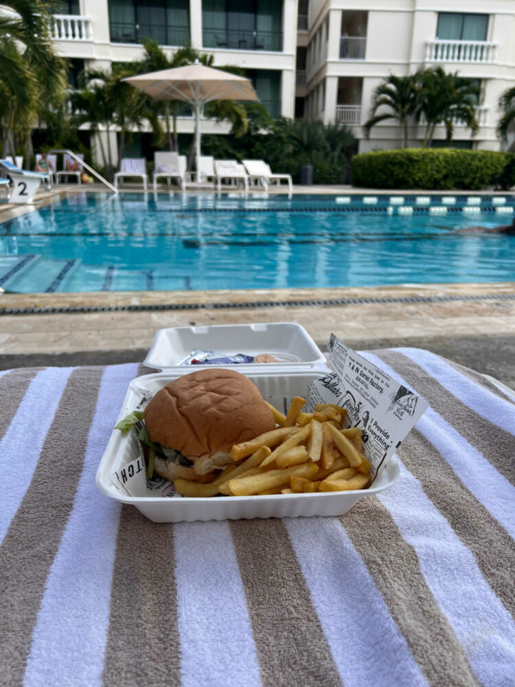 a hamburger and french fries in a takeout container on a striped towel next to a swimming pool