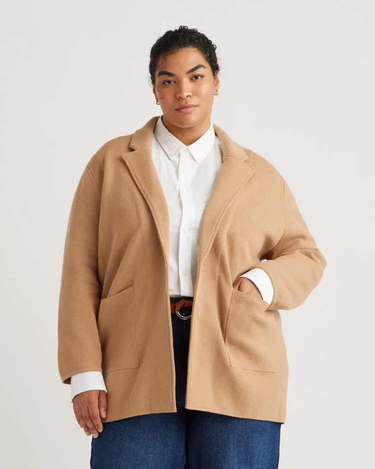 A model wearing thje camel Quince Organic Cotton Knit Blazer in Extended Sizes over a white shirt and jeans. One hand is in the blazer pocket.