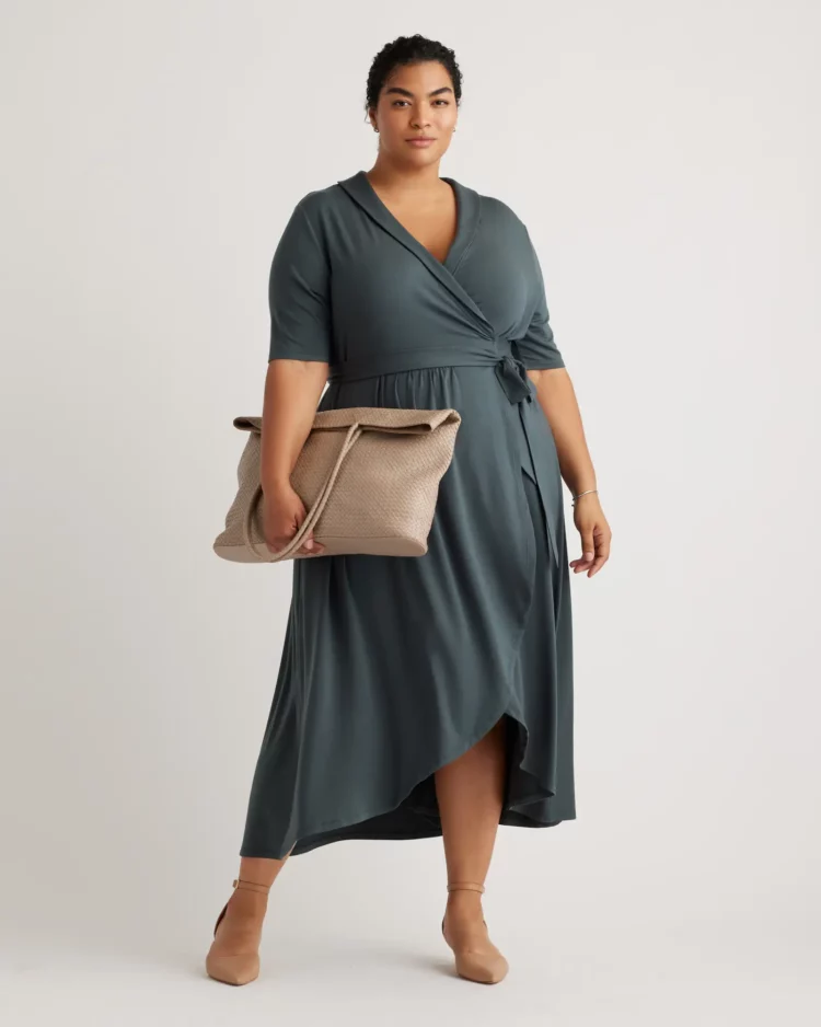Quince plus size model wearing the Quince tencel wrap dress in olive. She is holding a taupe handbag in one hand.