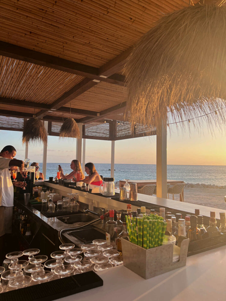 a view of a beachside bar in Curacao. Bartenders mixing drinks, two women sitting at the bar with cocktails, and the ocean behind them.