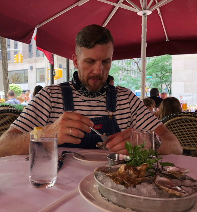 man in a J. Crew striped t-shirt and overalls with a bandana tied around his neck, sitting outside under a red umbrella eating raw oysters