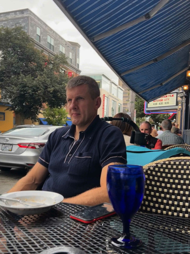 Man in a navy Todd Snyder Riviera polo shirt sitting at an outdoor cafe in Providence, Rhode Island. This is one of the best fashion buys for my husband I have made in the past decade.