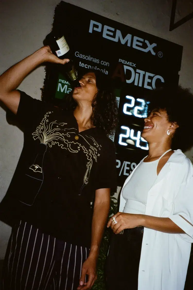 Man and woman in a club. The woman is looking at the man as he swigs champagne right from the bottle. He is wearing a black Tombolo cabana shirt of terrycloth with a champagne design embroidered on it.