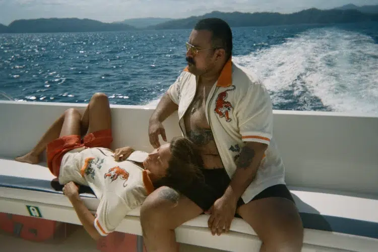 a man and a woman on a boat wearing matching zip front terrycloth Tombolo cabana shirts. The man is wearing his open with shorts. The woman has her head in the man's lap, wearing the shirt zipped up with red shorts