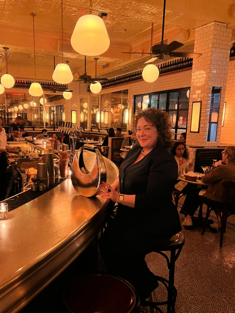 An over-40 woman with dark curly hair sitting at the bar at Pastis DC. She is waring a navy double breasted pantsuit and is holding a silver chrome STAUD moon bag. She is smiling at the camera, it is evening and dimly lit in the restaurant