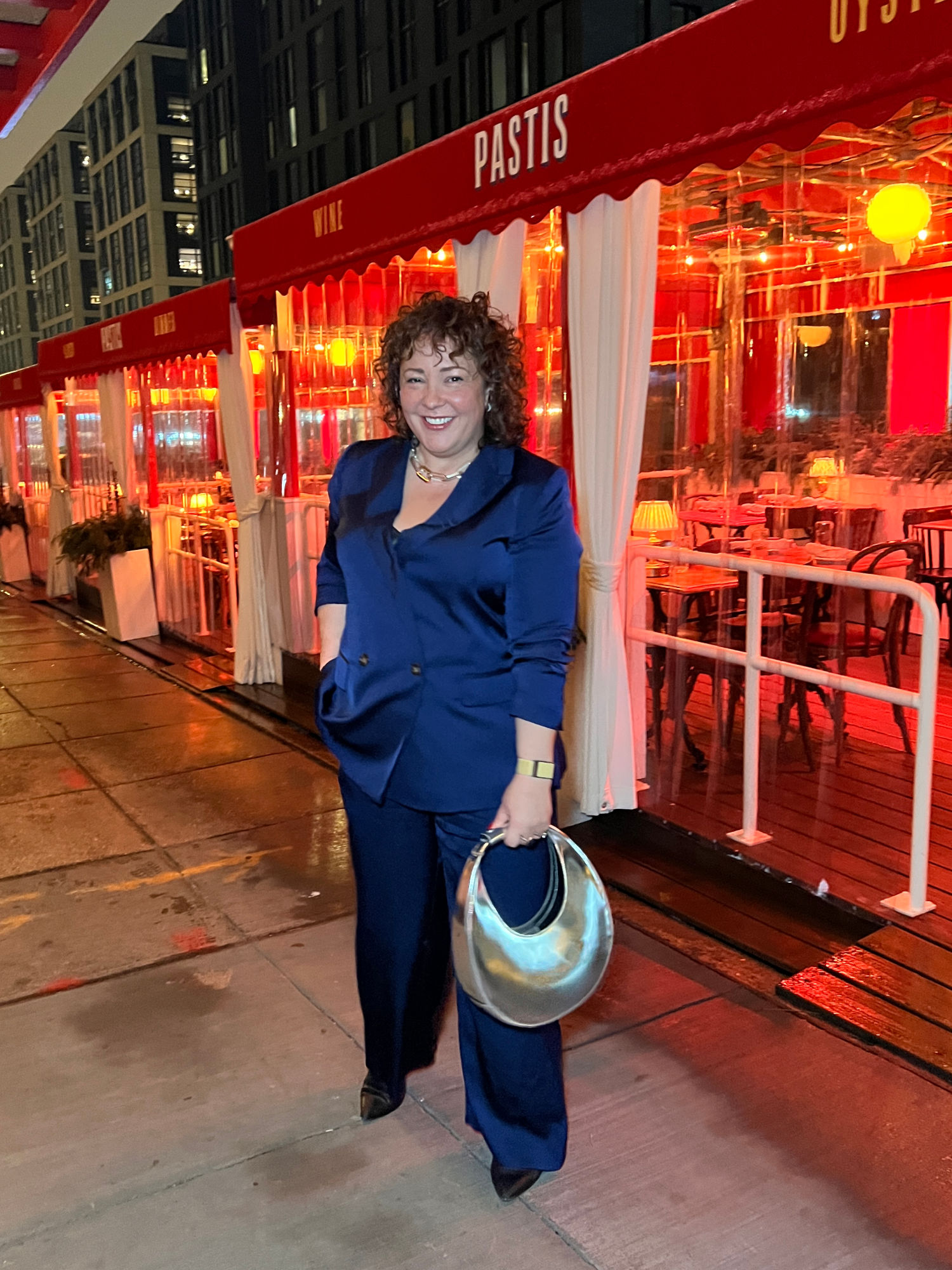 Alison Gary of Wardrobe Oxygen at Pastis DC. She is standing outside the restaurant in the Union Market District, in a shiny navy pantsuit, a silver bag in her hand. She has one hand in her pocket and she is smiling at the camera.