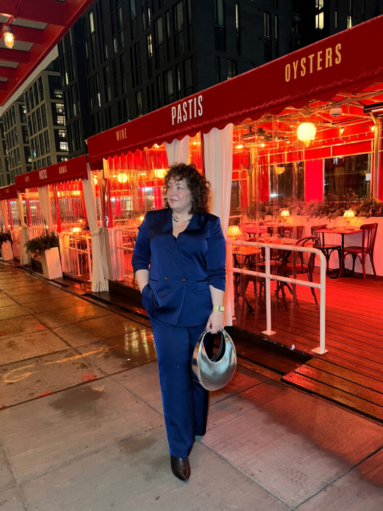 Alison Gary of Wardrobe Oxygen at Pastis DC. She is standing outside the restaurant in the Union Market District, in a shiny navy pantsuit, a silver bag in her hand. She has one hand in her pocket and she is smiling at the camera.