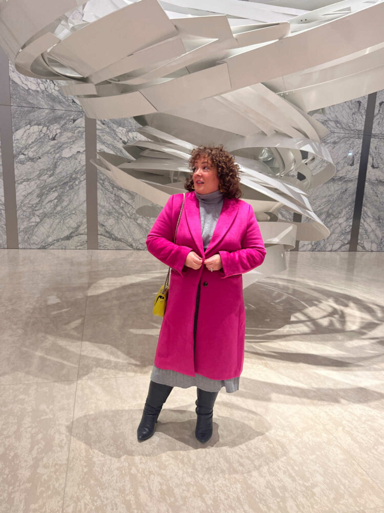Woman in a gray Quince cashmere turtleneck and matching pleated midi skirt. Over it she is wearing a hot pink wool below knee length coat. She has a pale lime green quilted Coach crossbody over it. On her feet are black knee high boots from Ros Hommerson. She is standing in front of a modern sculpture in the lobby of an office building.