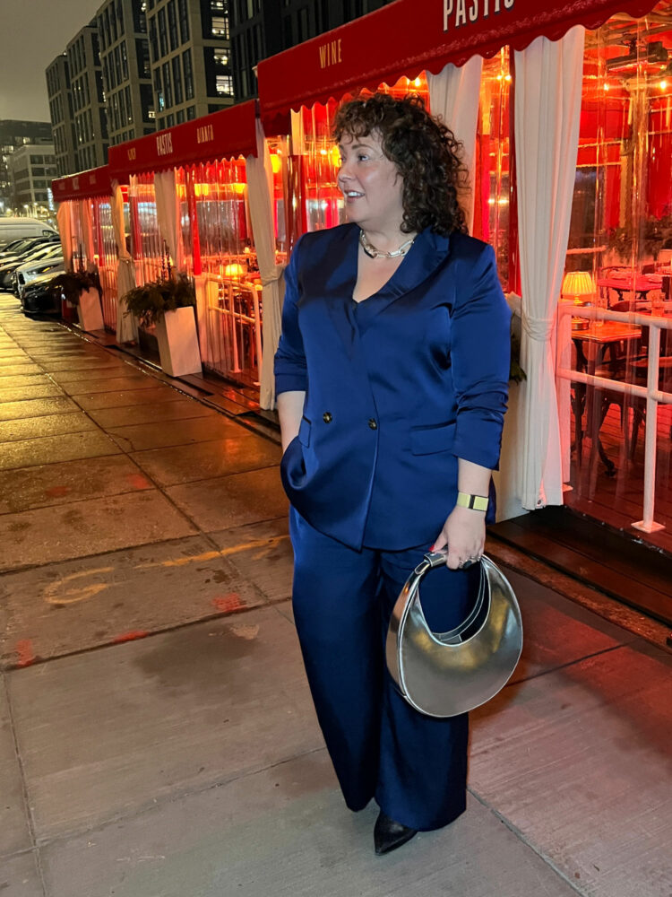Alison Gary, a petite size 14/16 woman over 40, is standing outside of Pastis DC in front of the outdoor dining area. She is in a navy satin double breasted pantsuit from Ann Taylor and is holding a shiny STAUD Moon bag in her hand. It is night and the sidewalk is damp from a recent rain.