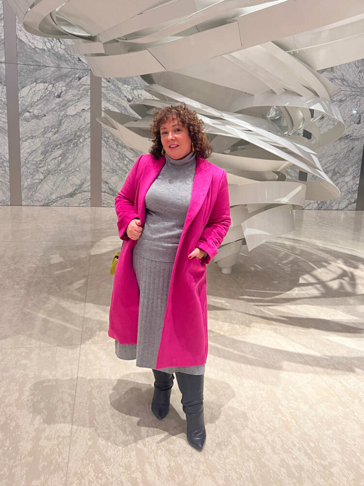 woman in a gray cashmere turtleneck and matching pleated midi skirt. She has a hot pink wool Chesterfield coat over it and has her had on her hip.