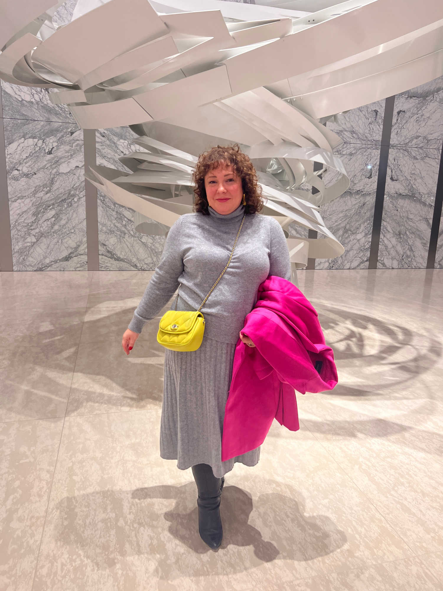 Alison of Wardrobe Oxygen in the lobby of an office building standing in front of a white modern art sculpture. She is wearing a gray cashmere turtleneck and matching skirt. She has a yellowish green crossbody bag and a hot pink wool coat in her hand. She is walking towards the camera.