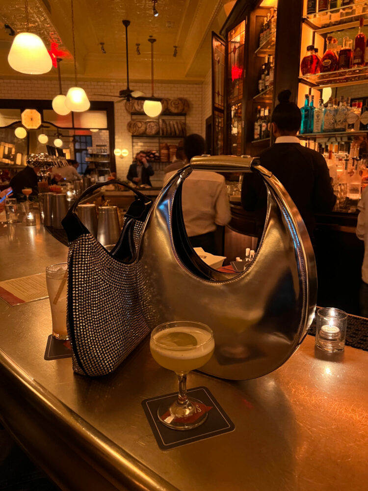 a chrome STAUD moon bag and a crystal-encrusted black crescent bag on the bar at Pastis DC with a cocktail in front called the Eiffel Sour