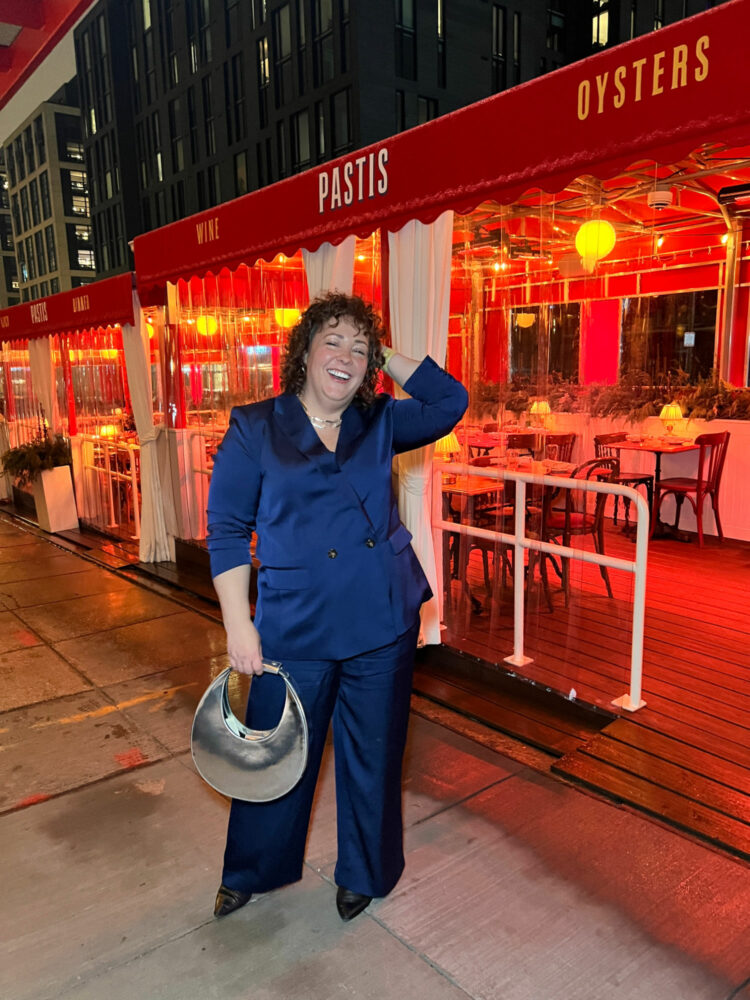 Alison Gary of Wardrobe Oxygen standing in front of the restaurant Pastis in the Union Market district of Washington DC. She is wearing a navy satin pantsuit. One hand is in her hair, the other is holding a silver chrome STAUD moon bag. She is laughing.