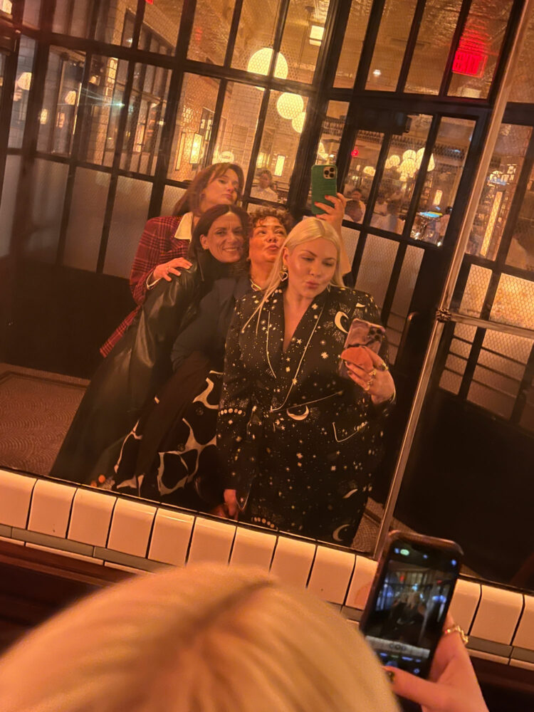 four women over 40 taking a mirror selfie in the entrance of Pastis DC. They are hugging each other; two are smiling and two are making kissy faces. They are fashionably dressed, it is evening.
