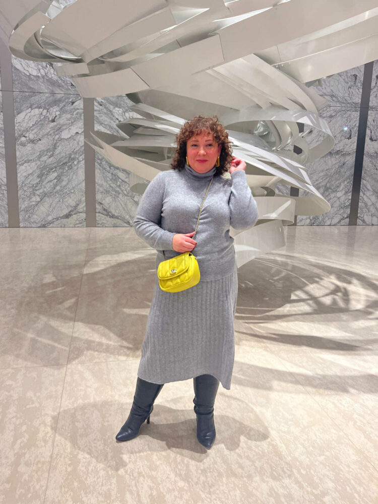 Woman with brown curly hair wearing a gray cashmere turtleneck and matching cashmere midi skirt. She has a light green crossbody bag with a chain strap and is wearing black knee high heeled boots