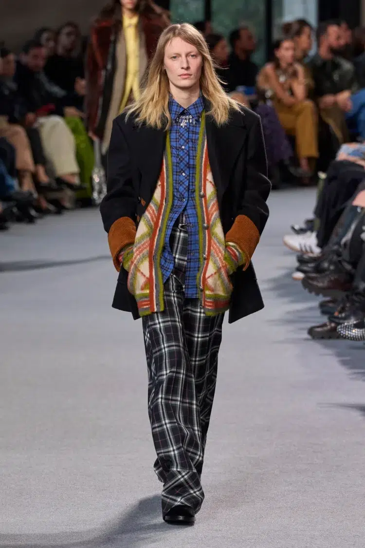 a look from Rabanne's Fall 2024 ready to wear collection of a model wearing plaid pants, a denim shirt, an argyle cardigan, and a coat over it all