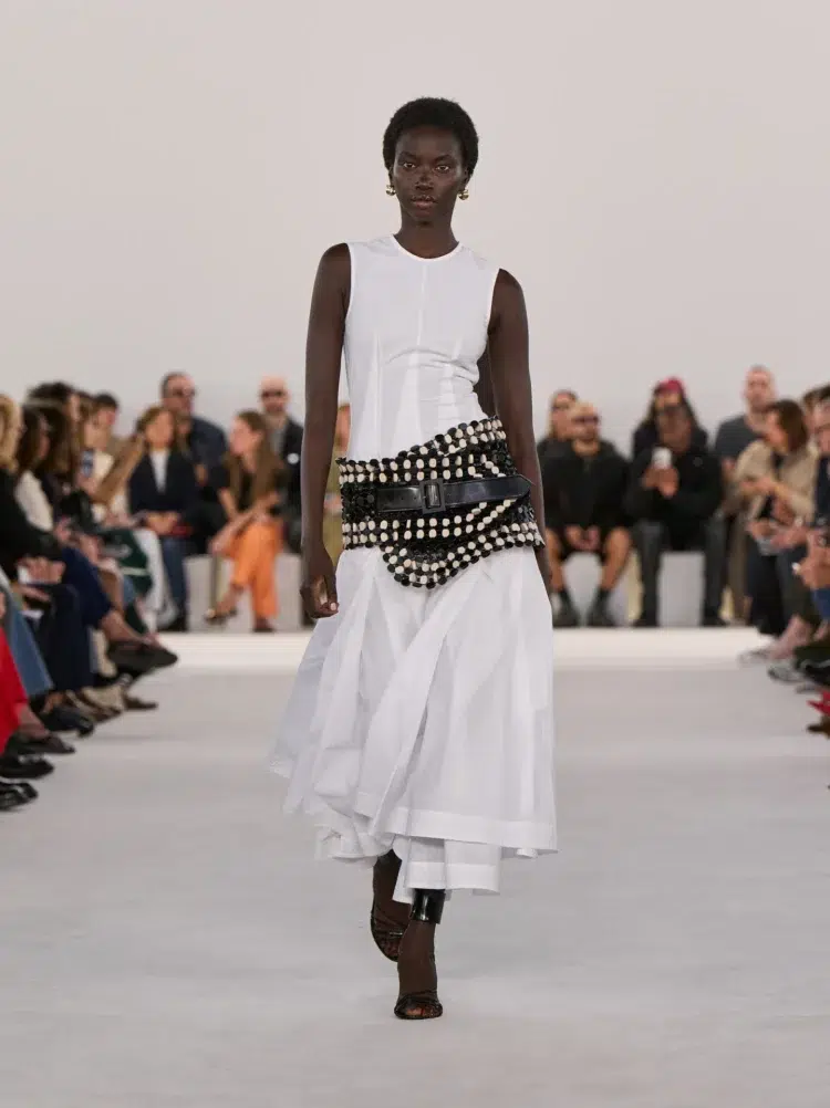 A white sleeveless fit and flare midi dress with a large black leather studded hip belt on a model at the Ferragamo S/S 2024 runway show