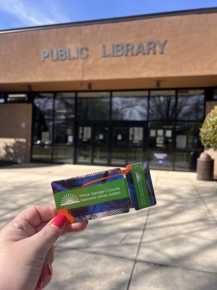 a woman's hand hoping up a Prince George's County library card in front of the Greenbelt Public Library in Greenbelt, Maryland