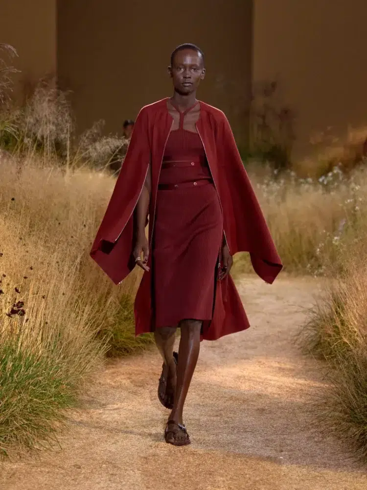 A look from Hermes Spring Summer 2024 runway show of a model in a dark red dress and matching cape coat hybrid.