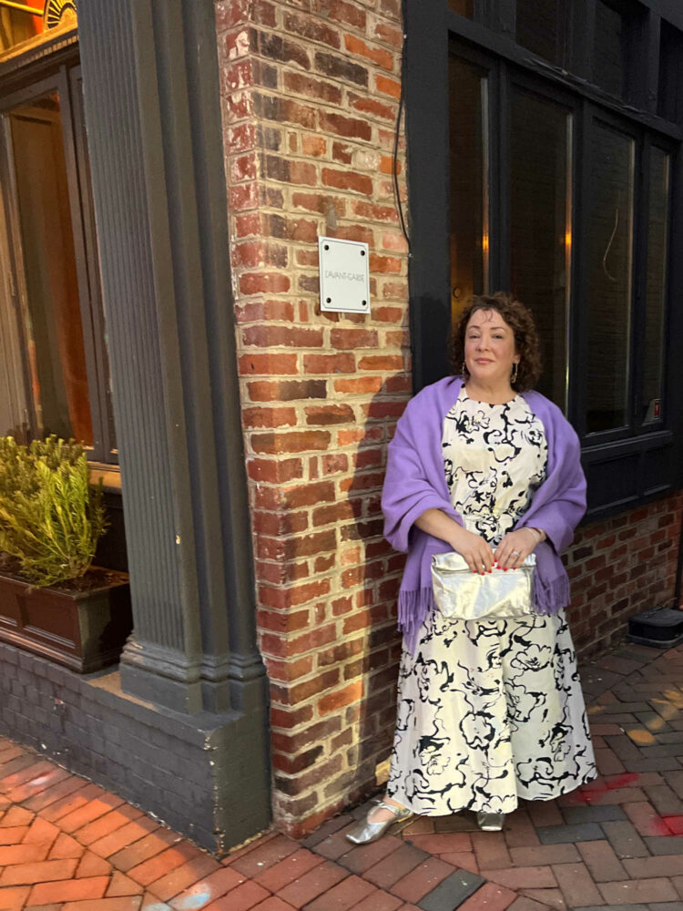 Alison Gary, a petite over-40 woman with curly brown hair is standing outside L'Avant-Garde in Georgetown. She is wearing a black and white patterned puff sleeve top and ankle length skirt. A lavender pashima is over her shoulders and she is holding a silver clutch purse.