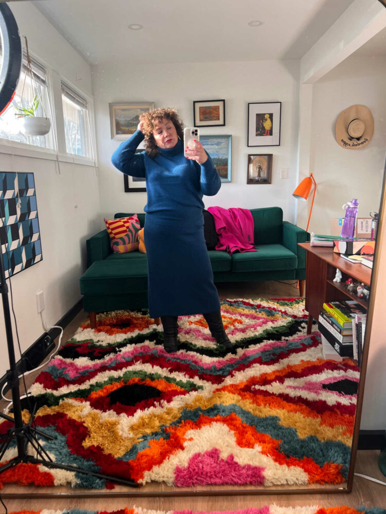 Alison Gary of Wardrobe Oxygen standing in her office on a multicolored shag rug. Behind her is a green velvet sofa; she is taking a selfie in a large gold framed mirror
