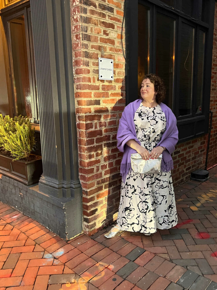 Alison Gary, a petite over-40 woman with curly brown hair is standing outside L'Avant-Garde in Georgetown. She is wearing a black and white patterned puff sleeve top and ankle length skirt. A lavender pashima is over her shoulders and she is holding a silver clutch purse.