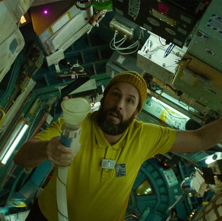Adam Sandler wielding his space urinal. No really, he is in a yellow t-shirt and knit cap floating in a spaceship holding a urinal with a tube looking confused.