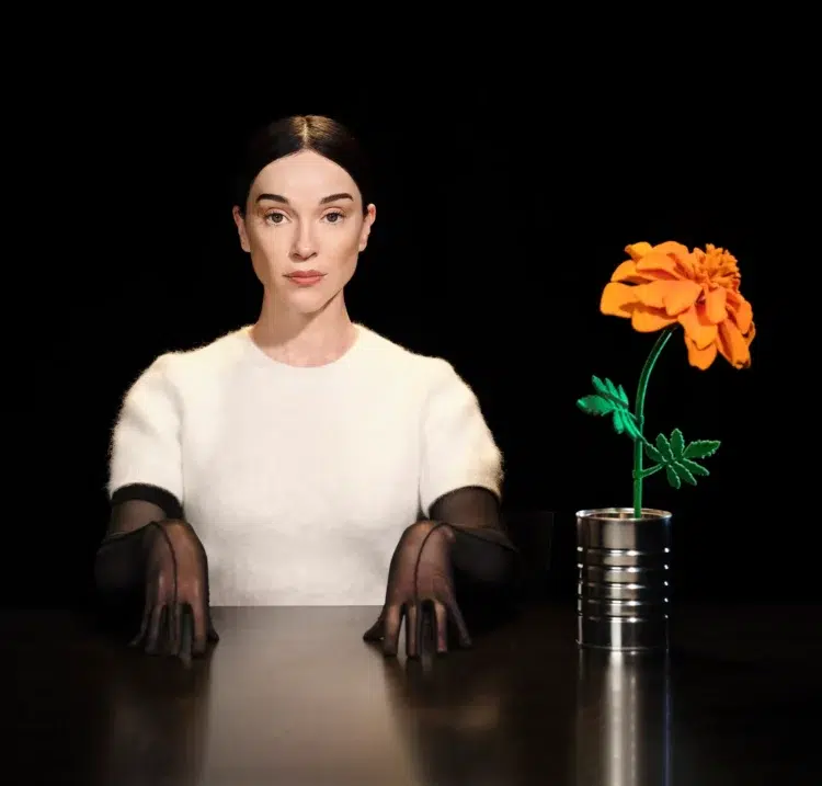 Annie Clark as St. Vincent. She is sitting at a table in a white short-sleeve top and black sheer gloves and ont he table next to her is a vase holding an orange flower. She is staring at the camera.