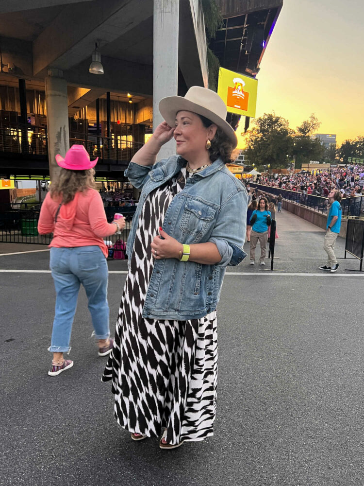 Woman at an outdoor concert in a brown and white graphic print silk trapeeze maxi dress, a cream felt western hat and the Universal Standard denim chore jacket