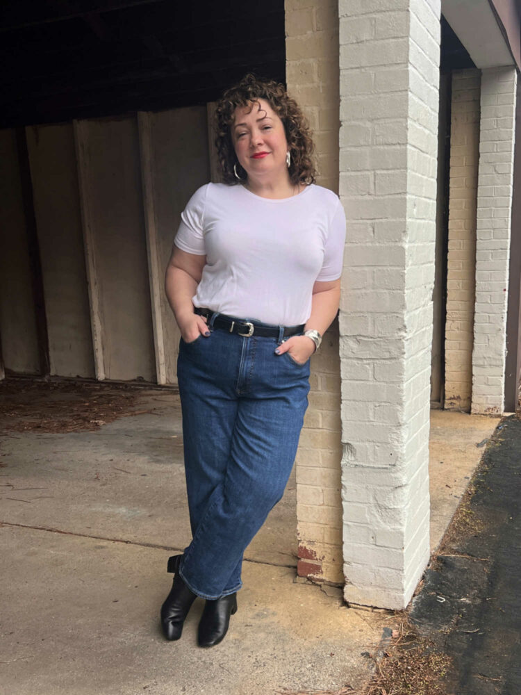 49 year old woman in a white crewneck t-shirt and medium wash straight leg jeans. She has on a black leather western belt and black boots and is leaning against a brick pillar