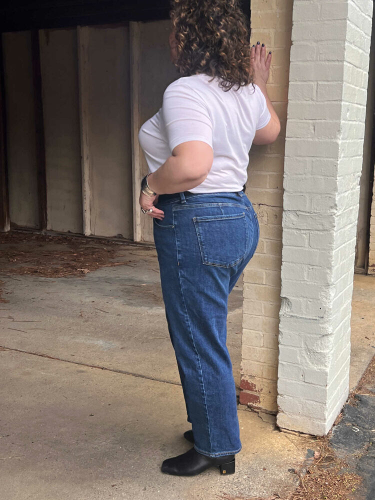 49 year old woman in a white crewneck t-shirt and medium wash straight leg jeans. She has on a black leather western belt and black boots and is leaning against a brick pillar