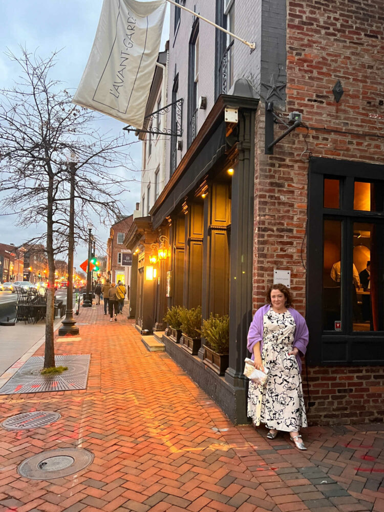 Alison Gary, a petite over-40 woman with curly brown hair is standing outside L'Avant-Garde in Georgetown. She is wearing a black and white patterned puff sleeve top and ankle length skirt. A lavender pashima is over her shoulders and she is holding a silver clutch purse.