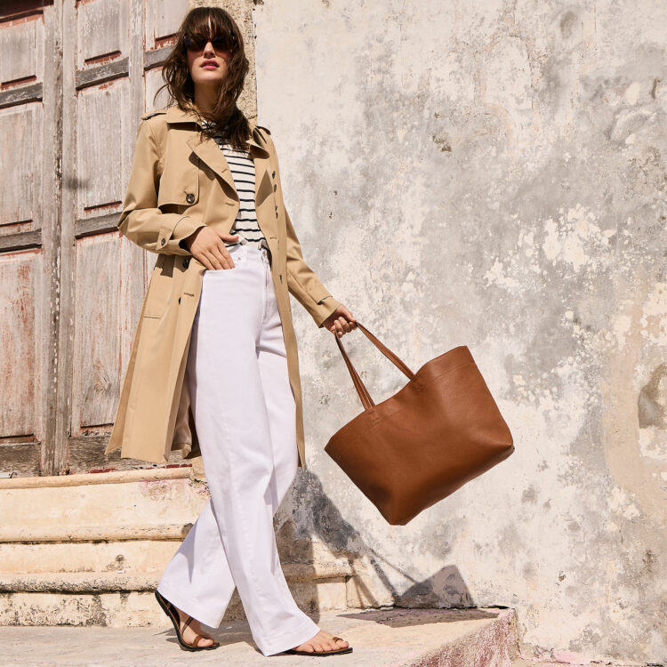 a look from Lands' End spring collection of a model in a Breton striped top, white wide leg chinos, a trench, anda tan leather tote walking outside.