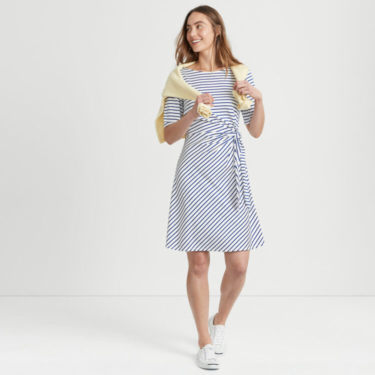 a model for Lands' End wearing a blue and white striped fit and flare dress with white sneakers and a yellow sweater around her shoulders