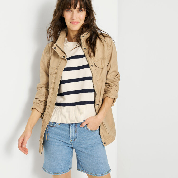 Lands' End model in a tan twill jacket, cream sweater with navy stripes, and knee-length faded denim shorts