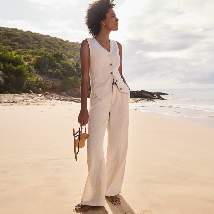 A woman on a beach wearing Lands' Ed spring linen vest and matching pants, looking at the water, a pair of sandals in her hand