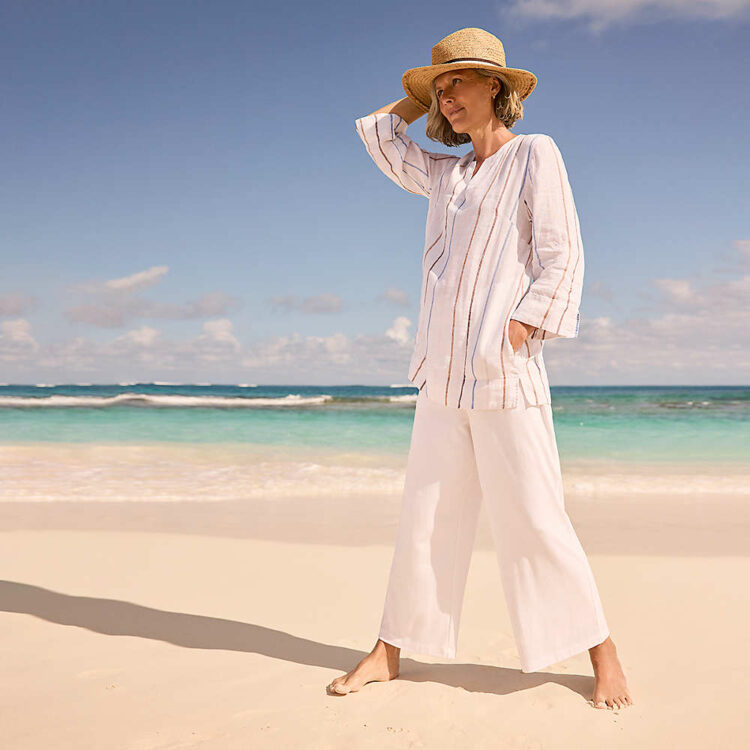 A woman in a white striped linen tunic and white wide leg pants on a beach holding a straw panama hat on her head