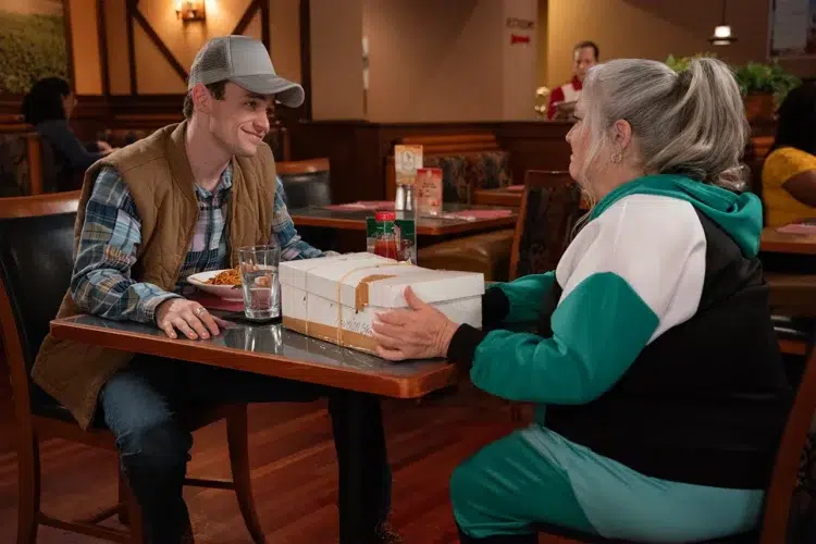 Paula Pell with the character Mad Dog sitting in a restaurant in Season 3 of Girls5Eva.