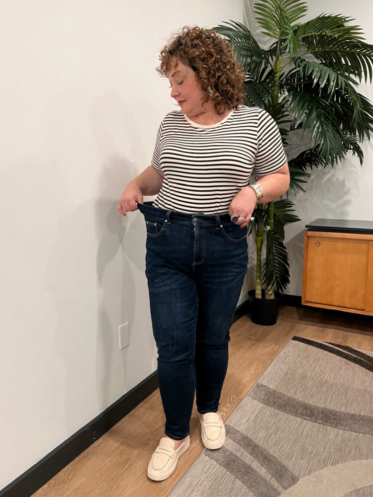 Alison wearing a striped t-shirt tucked into Universal Standard Joni jeans in a dark wash. She is pulling the waistband to the side to show the stretchiness of the jeans.
